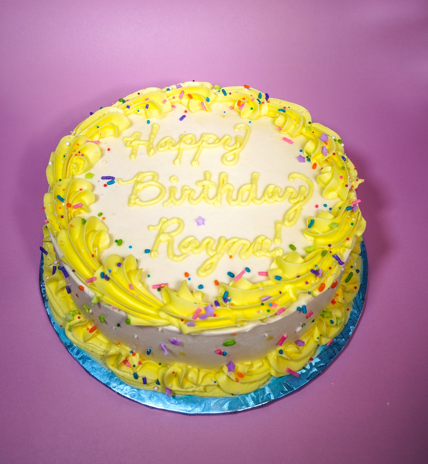 A cake frosted white with a yellow border and yellow writing that says, "Happy Birthday Rayna!"