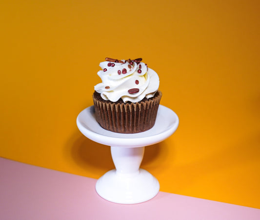 A chocolate cupcake with white frosting on a white cupcake stand in front of an orange and pink background